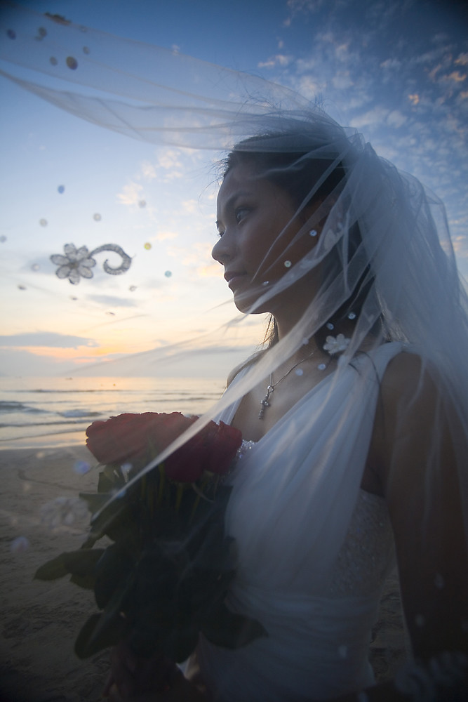 Noiva de véu na praia durante o pôr do sol, com o mar ao fundo.