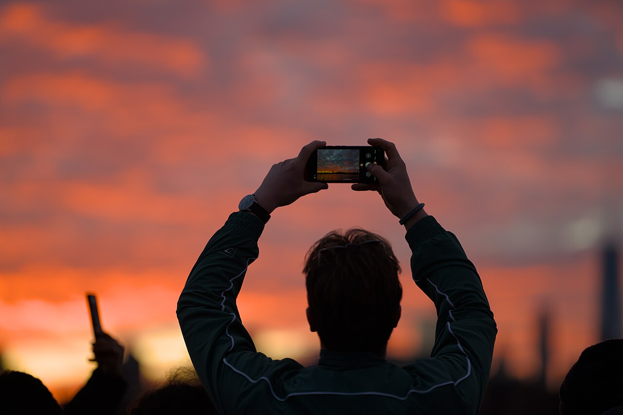 Pessoa de costas segurando o celular na horizontal para fotografar o céu alaranjado do pôr do sol.