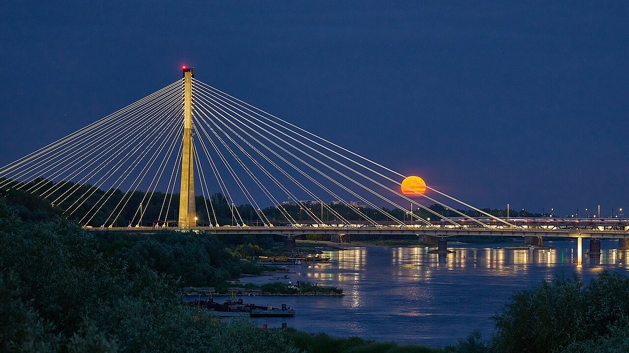 Lua cheia alaranjada surgindo no horizonte atrás de uma ponte estaiada, ao anoitecer, refletida no rio