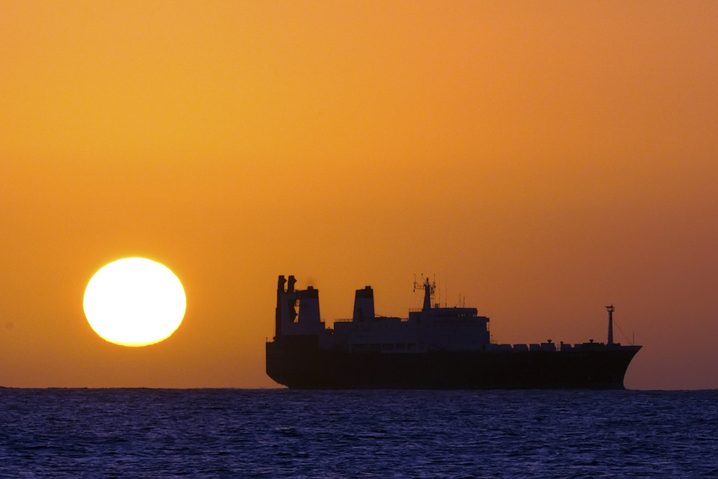 Sol baixo sobre o oceano ao amanhecer, com um navio em silhueta no horizonte.