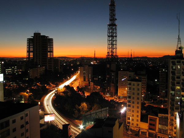 Skyline de São Paulo ao entardecer, com o horizonte alaranjado atrás dos prédios e as avenidas já acesas.