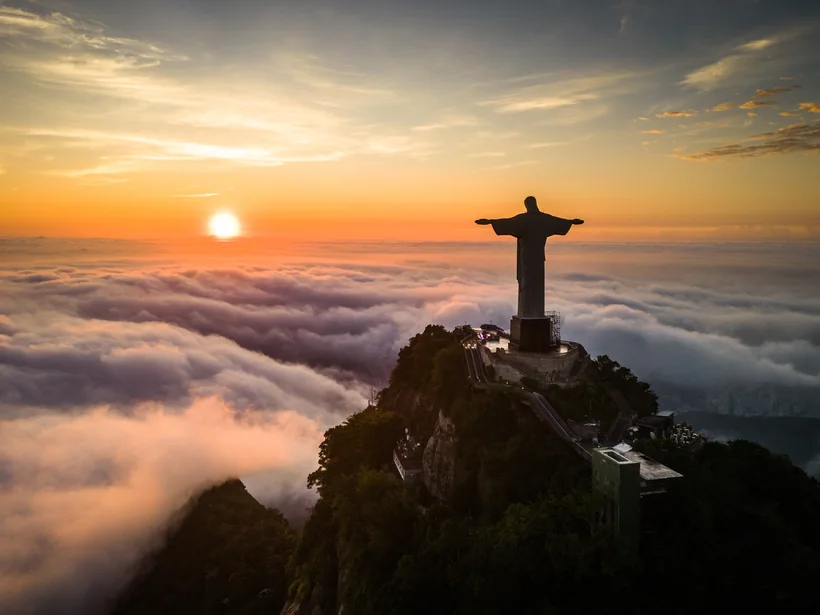 Estátua do Cristo Redentor no topo do Corcovado ao pôr do sol, acima de um mar de nuvens